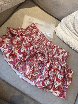 Ny i förpackning! Röd paisleymönstrad volangkjol - Supersöt kort kjol med rött och vitt paisleymönster och bred smock i midjan. Kjolen har volangdetaljer och är perfekt för dig som gillar en lekfull och trendig stil. Lätt och luftig känsla, passar perfekt till varma dagar.