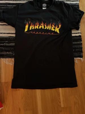 Svart Thrasher t-shirt med flames - Svart t-shirt från Thrasher med ikoniskt flames-tryck och gul logga på bröstet. Klassisk passform och rund halsringning. Perfekt för dig som gillar streetwear och skatekultur.