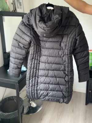 Svart lång dunjacka med huva - Snygg svart dunjacka i längre modell med huva och två dragkedjeförsedda fickor framtill. Jackan har quiltad design och dragkedja hela vägen upp. Perfekt för kalla vinterdagar och håller dig varm och stilren.