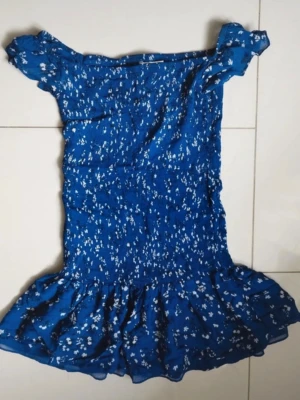  Ny slutsåld Blå blommig kortklänning från Zara - Blå kortklänning från Zara med vita blommor och volang nertill. Klänningen har offshoulder-design och är tillverkad i ett lätt och luftigt material, perfekt för varma dagar. Snygg och trendig stil som passar dig som gillar att sticka ut.