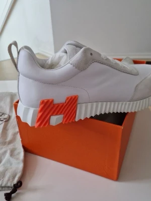 Vita H skor - Säljer ett par vita H sneakers med grymt cool orange H-detalj på sidan av sulan. Skorna har en chunky, räfflad sula och ovandel i läder och mocka. Perfekta för dig som vill sticka ut med stilrena men unika sneakers.