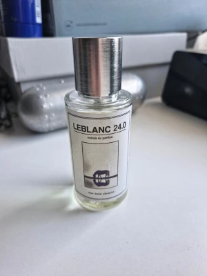 LEBLANC 24.0 Extrait de Parfum - LEBLANC 24.0 Extrait de Parfum i en stilren, genomskinlig glasflaska med metallfärgad kork. Parfymen har en ljusgul vätska och etiketten pryds av en minimalistisk design med text och en illustration. Perfekt för dig som gillar unika dofter. Är från oneboldchemist är i bra skick