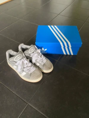 Grå Adidas sneakers i mocka - Snygga grå sneakers från Adidas med klassiska vita ränder på sidorna och breda vita skosnören. Skorna är låga och har en ovandel i mocka samt Adidas logga på plösen. Yttersulan är i gummi med beige ton. Perfekta för en avslappnad streetstyle.