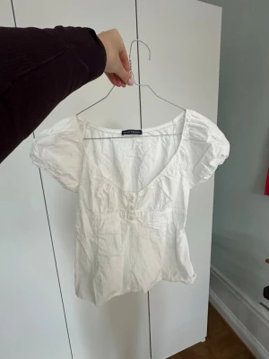 Vit topp med puffärmar från brandy Melville - Söt vit topp  med puffärmar och knappar framtill. Modellen har en djup rundad urringning och smock i midjan för en snygg passform. Tillverkad i ett lätt bomullstyg som känns luftigt och fräscht.