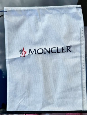 Vit tunn dustbag från Moncler - Vit dustbag från Moncler med dragsko upptill och Moncler-logga i svart och rött framtill. Tillverkad i ett tunt tygmaterial, perfekt för att förvara väskor, skor eller accessoarer. 