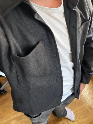 Mörkgrå overshirt med ficka - Snygg mörkgrå overshirt med klassisk krage och knappstängning framtill. Overshirten har en stor bröstficka och är gjord i ett tjockare tyg som ger en stilren och avslappnad look. Perfekt att bära över en t-shirt för en casual vibe.