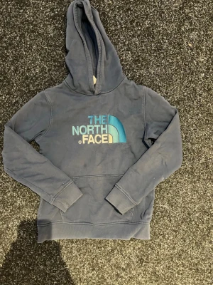 Blå hoodie från The North Face - Mörkblå hoodie från The North Face med stor logga i blått och vitt på bröstet. Klassisk känguruficka framtill, ribbade muddar och huva. Tillverkad i mjukt bomullsmaterial som är skönt att ha på sig. Perfekt för en avslappnad och cool stil. Kolla gärna in mina andra annonser 