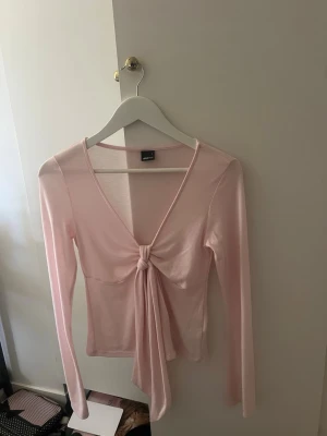 Ljusrosa långärmad topp med knyt - Trendig ljusrosa topp med lång ärm och djup v-ringning. Toppen har en snygg knyt-detalj framtill som ger en feminin touch. Tillverkad i mjukt och stretchigt material som sitter bekvämt på kroppen. Perfekt för att lyfta vilken outfit som helst.