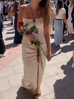 Champagnefärgad långklänning satin - Stilren långklänning i champagnefärgad satin med smala axelband och en snygg slits framtill. Klänningen har en figurnära passform och minimalistisk design som ger en lyxig känsla. Perfekt till student eller bal.