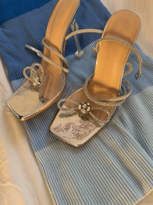 Silvriga pumps med strassdetaljer - Glammiga silvriga pumps med fyrkantig tå och transparent klack. Skorna har tunna remmar dekorerade med strass och en gnistrande blomdetalj framtill. Perfekta för dig som vill sticka ut och addera extra bling till din outfit.