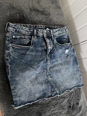 Blå jeanskjol - Säljer en blå jeanskjol i klassisk denim med rå, fransig kant nertill och slitning på framsidan. Kjolen har fickor, bälteshällor och dragkedja med knapp. Perfekt för en avslappnad och trendig look. Blev förvånad över storleken då jag alltid trott att den är i XS/S, jag har använt den trots dens storlek och då är jag ändå 17 och 1,65😌