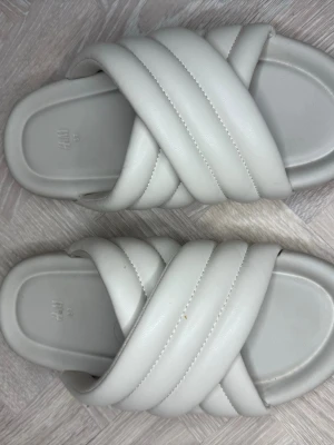 Vita vadderade sandaler från H&M - Säljer ett par trendiga vita sandaler från H&M med breda, korslagda och vadderade remmar. De har en chunky sula och är tillverkade i ett mjukt, konstläderliknande material. Perfekta för en avslappnad och stilren look.