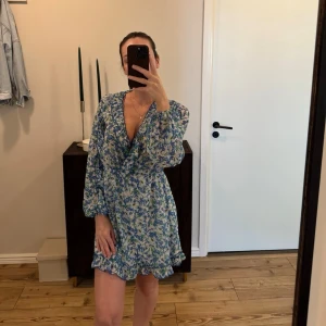 Blommig klänning i blått och vitt - Oanvänd, söt kort klänning med blått och vitt blommönster. Klänningen har volangkant nertill, v-ringning och ballongärmar. Perfekt för sommarens alla tillfällen och ger en romantisk look.