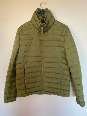 Dunjacka  - Säljer en olivgrön dunjacka från Dressmann med hög krage och dragkedja framtill. Jackan har quiltad design och praktiska fickor både på utsidan och insidan. Perfekt för kyliga dagar och riktigt snygg till jeans eller cargo.Den är som ny.