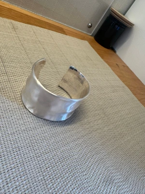 Brett silverfärgat metallarmband - Snyggt och brett armband i silverfärgad metall med en lätt vågig och hamrad yta. Öppen design som gör det enkelt att ta på och av. Inte använt så mycket. 