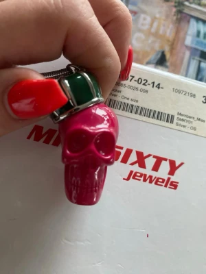 Miss Sixty halsband med dödskalle - Unikt halsband från Miss Sixty med en stor röd dödskalle som hänge med krona. Kedjan är i silverfärg och har en liten rund bricka med Miss Sixty-loggan. Perfekt statement piece för dig som gillar edgy accessoarer. Box certifikat ingår~ vintage!