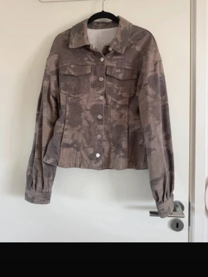 Mönstrad jeansjacka - Säljer en khaki jeansjacka med coolt tvättat camo-mönster. Jackan har klassisk krage, två bröstfickor med lock och silverfärgade knappar framtill. Modellen är något kortare och har långa ärmar med knapp vid ärmslut. Perfekt för dig som vill sticka ut lite extra.