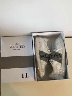 Valentino Garavani vita sneakers - Snygga vita sneakers från Valentino Garavani med en bred grå rem över sidan och klassisk låg siluett. Skorna har rund tå, vita snören och diskreta nitar på hälen. Perfekta för dig som vill ha en clean men ändå lyxig look. Jag gör såklart rent skorna innan jag skickar dem!!