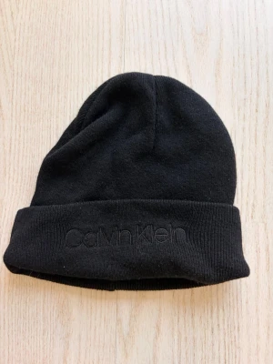 Svart mössa från Calvin Klein - Säljer en svart ribbstickad mössa från Calvin Klein med uppvikt kant och diskret broderad logga framtill. Klassisk och enkel design som passar till det mesta. Tillverkad i mjukt material som håller dig varm under kalla dagar.