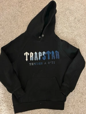 Trapstar hoodie äkta - Svart hoodie från Trapstar med stor huva och magficka. På bröstet står det 'TRAPSTAR' i gradient från vitt till blått och texten 'IT'S A SECRET' under. Klassisk streetwear-look med ribbade muddar och mjukt material, perfekt för dig som gillar en avslappnad stil.