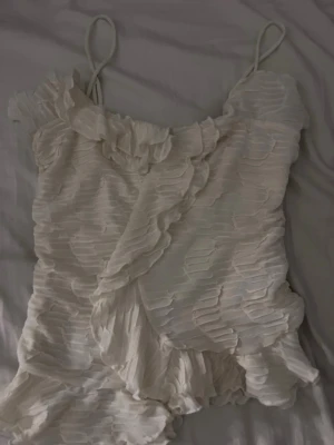 Vit omlott linnetopp med volang - Säljer en vit linnetopp med tunna axelband och snygga volangdetaljer. Toppen har ett omlott-design och ett strukturerat mönster som ger en cool look. Perfekt för dig som gillar unika och trendiga plagg med lite extra detaljer.