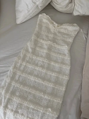Vit spetsklänning med omlott - Säljer en vit långklänning med vacker spets och omlott-detaljer. Klänningen är ärmlös och har en feminin v-ringning. Perfekt för dig som vill ha en romantisk och elegant look. Materialet är troligtvis bomull eller polyester med spets.