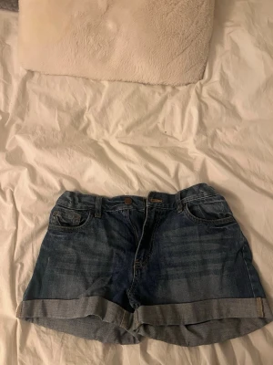 Blå jeansshorts med upprullad kant - Snygga blå jeansshorts med upprullad kant och klassisk femficksdesign. Shortsen har bälteshällor, knapp och dragkedja framtill samt två bakfickor. Perfekta för varma dagar och enkel att matcha med olika stilar.