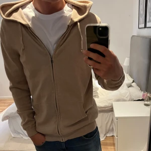 Beige hoodie med dragkedja från Tresanti - Snygg beige hoodie från Tresanti med dragkedja framtill, huva med snörning och två fickor. Tröjan är i mjuk bomullskvalitet samt Kashmir och har ribbade muddar vid ärmslut och nederkant. Perfekt för en chill och avslappnad stil. Tveka inte  att fråga vid funderingar!