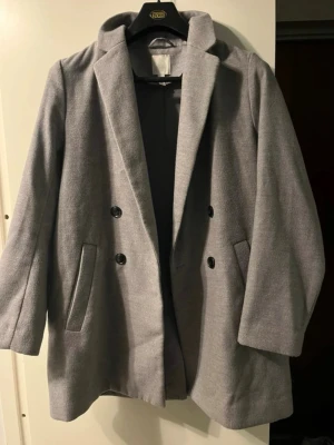 Ljusgrå dubbelknäppt kappa - Stilren ljusgrå kappa med dubbelknäppning och två framfickor. Klassisk krage och rak passform ger en clean look. Perfekt för kyligare dagar och enkel att matcha med olika outfits.  Endast använd några gånger så i fint skick ☀️