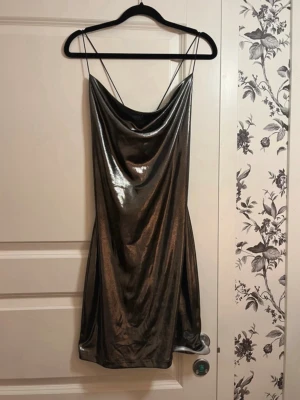 Glittrig silver kort klänning - Säljer en glittrig silverfärgad kort klänning med tunna axelband som korsas i ryggen. Klänningen har en snygg draperad urringning och är tillverkad i ett glansigt material som ger en festlig look. Använd vid ett tillfälle :)