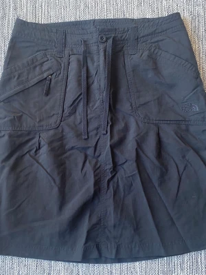 Svart kjol från The North Face - Snygg svart kjol från The North Face med knytband i midjan och stora fickor fram. Tillverkad i ett slitstarkt material som passar perfekt för vardagsäventyr. Diskret logga på ena fickan och knappstängning i midjan. Svart friluftskjol i strl S från The North Face  Använd men i fint begagnat skick   Mått Midjan: ca 41 cm Total längd: ca 54 cm  100% nylon  Kommer från ett djur och rökfritt hem. 