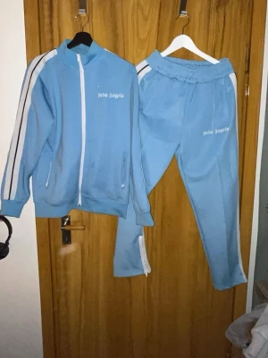 Palm Angels tracksuit - Säljer ett ljusblått tracksuit-set från Palm Angels med vita och svarta ränder längs ärmar och ben. Setet består av en zip-jacka med hög krage och två fickor samt matchande byxor med resår i midjan. Båda delarna har Palm Angels-loggan tryckt i vitt.