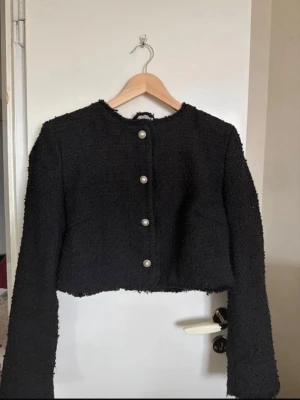 Svart cropped kofta med pärlknappar - Säljer en svart cropped kofta i bouclé-liknande material med fyra dekorativa pärlknappar framtill. Rund halsringning för en trendig look. Långa ärmar och figurnära passform. 