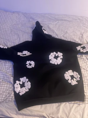 Svart hoodie från Denim Tears - Säljer en svart hoodie från Denim Tears med stora vita blomtryck över hela tröjan. Hoodien har en klassisk huva och är gjord i ett mjukt och bekvämt material, perfekt för en avslappnad streetwear-look.