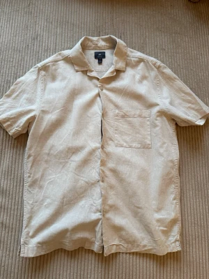 Beige linneskjorta från H&M - Säljer en stilren beige skjorta från H&M i linne med korta ärmar och bröstficka. Skjortan har klassisk krage, knappar framtill och en avslappnad regular fit. Perfekt för varma dagar och enkel att matcha med jeans eller shorts.