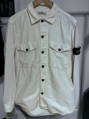 Vit overshirt från Stone Island - Säljer en stilren vit overshirt från Stone Island med klassisk krage, svarta knappar och två bröstfickor med lock. Ikonisk Stone Island patch på vänster ärm. Tillverkad i ett mjukt bomullsmaterial som ger en clean och modern look. 100 procent äkta. Pris kan absolut diskuteras 