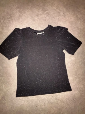 Svart glittrig topp med puffärm - Snygg svart topp med glittrigt tyg och puffiga korta ärmar. Perfekt för dig som vill sticka ut lite extra. Rund halsringning och figurnära passform. Enkel att matcha med både jeans och kjol.