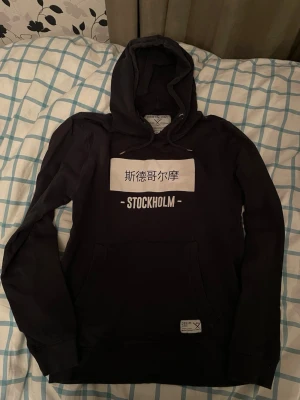Svart hoodie från Denim Day S - Svart hoodie från Denim Day med huva och stor ficka framtill. Tryck på bröstet med kinesiska tecken och texten STOCKHOLM. Mjuk bomullskänsla och avslappnad passform, perfekt för en chill look.
