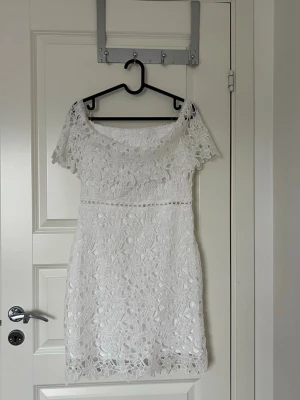 Studentklänning  - Kort vit klänning med vackert blommigt spetsmönster och offshoulder-design. Klänningen har korta ärmar och en feminin, figurnära passform. Perfekt för sommarens alla tillfällen där du vill känna dig extra fin.