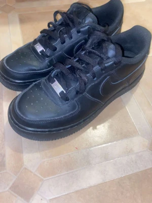 Svarta Nike Air Force 1 sneakers - Klassiska helsvarta Nike Air Force 1 sneakers i läder med svart sula och svarta skosnören. Ikonisk swoosh på sidan och metallbricka på snörningen. Perfekta för dig som gillar en clean och tidlös look.