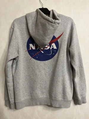 Grå NASA hoodie med logga - Snygg grå hoodie med NASA-logga tryckt både fram och stort på ryggen. Klassisk känguruficka och dragsko i huvan. Tillverkad i mjukt sweatshirtmaterial, perfekt för en avslappnad och cool stil.