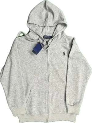 Grå zipper Polo Ralph Lauren - Snygg ljusgrå hoodie med dragkedja från Polo Ralph Lauren. Klassisk design med huva, två fickor framtill och den ikoniska lilla loggan broderad på bröstet. Tillverkad i mjukt bomullsmaterial som är perfekt för chill dagar.