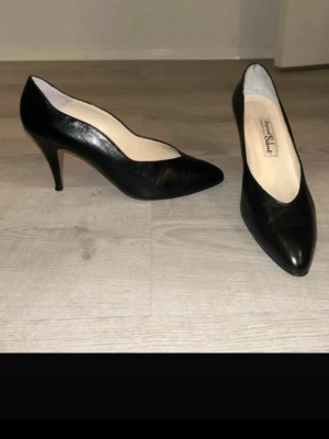 Svarta pumps från Salvatore Ferragamo - Klassiska svarta pumps från Salvatore Ferragamo i elegant läder. Skorna har spetsig tå och smal klack som ger en tidlös och stilren look. Insidan är ljus och mjuk för extra komfort. Perfekta för dig som gillar en chic och sofistikerad stil.