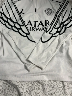  PSG träningströja 2026 - Säljer en vit långärmad träningströja från PSG med svart vingmönster på ärmar och bröst. Tröjan har halv dragkedja, PSG-logga på bröstet och Qatar Airways-tryck framtill. Tillverkad i ett lätt och tekniskt material som passar perfekt till träning.