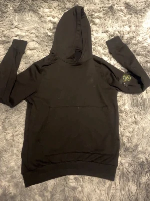 Svart hoodie från Stone Island - Säljer en svart hoodie från Stone Island med klassisk patch på vänster ärm. Hoodien har stor huva och magficka framtill. Tillverkad i mjukt bomullsmaterial och har ribbade muddar vid ärmslut och nederkant. Perfekt för en avslappnad streetwear-look.