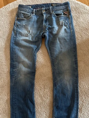 Blå slitna jeans från Replay - Säljer ett par blå jeans från Replay med slitningar och distressed detaljer på framsidan. Klassisk femficksmodell med raka ben och läderpatch bak i midjan. Jeansen har en snygg tvättad look och är tillverkade i denim. Str 32,32