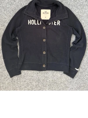 Mörkblå kofta från Hollister - Snygg mörkblå kofta från Hollister med knappar framtill och vit logotyp på bröstet. Koftan har hög krage och långa ärmar, samt diskret Hollister-märke på ärmen. Perfekt för att skapa en avslappnad och cool look. M men sitter som xs