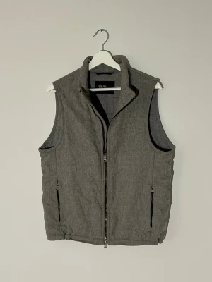 Grå väst från Oscar Jacobson - Stilren grå dunväst från Oscar Jacobson med hög krage och två dragkedjeförsedda fickor framtill. Västen har en rak passform och diskreta sömmar baktill. Perfekt att slänga över en hoodie eller tröja för extra värme.