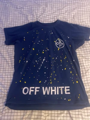 Blå Off White t-shirt med tryck - Snygg blå t-shirt från Off White med vitt och gult stänkmönster. Stor logga och text på både fram- och baksidan, samt grafiska detaljer och tryck som 'DO NOT CROSS' och 'TEMPERATURE'. Perfekt för dig som gillar streetwear och statement-plagg.