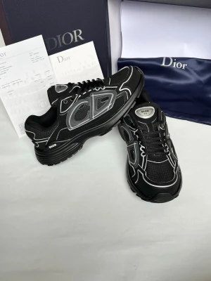 Svarta Dior sneakers med vita detaljer - Snygga svarta sneakers från Dior med vita och grå detaljer samt reflekterande partier. Skorna har en chunky sula, mesh- och läderpaneler samt tydlig Dior-logga på sidan. Perfekta för dig som vill ha en exklusiv och trendig look.